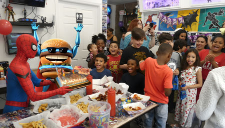Kids cheering at an indoor birthday party as a person in a Spider-Man costume and a smiling burger mascot present a lit birthday cake over a table of fries and party food