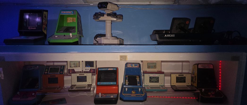 Retro gaming shelf display with vintage mini arcade cabinets and collectibles: tabletop arcade machines (including a green Frogger mini-cabinet), a white robotic console accessory, and Atari 2600 joysticks, lit by blue overhead light and red LED accents.