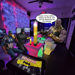 Neon-lit gaming lounge with glowing cloud-like LED wall, a Monopoly touchscreen table, and comic heroes Captain America, Batman and Thanos gathered around a tall glowing drink tower.