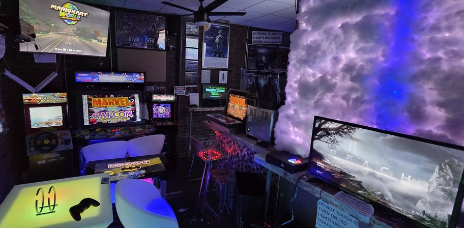 Neon-lit indoor arcade gaming lounge with retro cabinets (Marvel vs. Capcom), large TVs showing a racing game, glowing cloud-like purple-blue wall, multiple monitors, illuminated tables and white seating for modern and classic gaming.