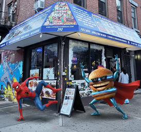 City corner burger shop with a bright blue awning and graffiti mural; Spider-Man crouches on the sidewalk beside a cartoon superhero cheeseburger mascot wearing sunglasses and a red cape, menu boards and city storefront visible.