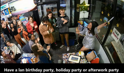 Crowded indoor LAN party celebration with people clapping around a table of cake and snacks while a woman in a sparkly dress leads cheering near a "LAN PARTY" sign and star balloon.