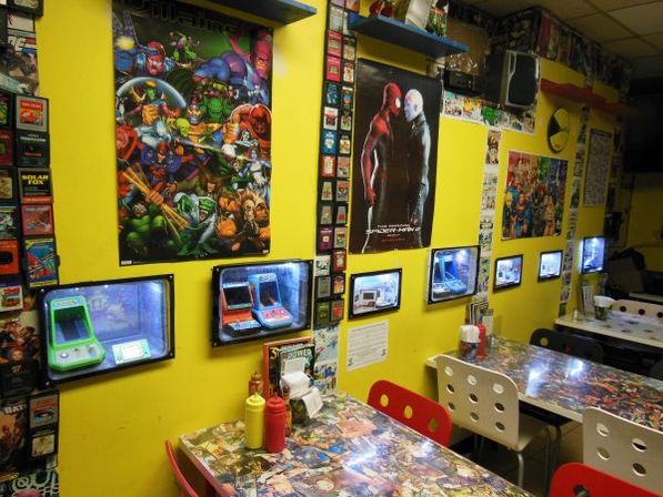 Retro arcade-style cafe interior with vibrant yellow walls, comic-book and superhero posters, built-in handheld game displays in glass wall niches, collage-covered tables, red and white chairs and ketchup and mustard bottles