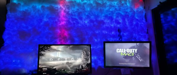 Home gaming room with dual monitors (Call of Duty MW3 and REACH) set before a blue‑purple LED cloud wall with starry ambient lighting