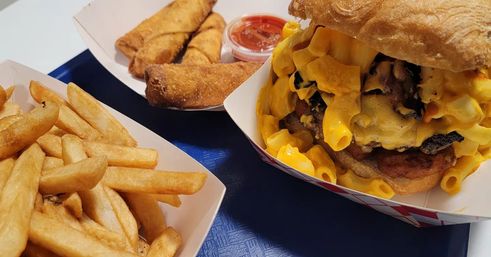 Close-up of a mac-and-cheese burger overflowing with creamy cheddar, golden fries and two crispy egg rolls with dipping sauce on a blue tray