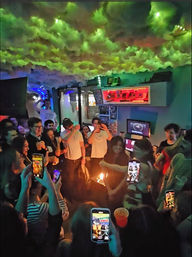 Vibrant urban nightlife scene: friends crowd a small bar for a birthday celebration, candles on a cake, phones recording under neon lights and a cloud-like ceiling installation.