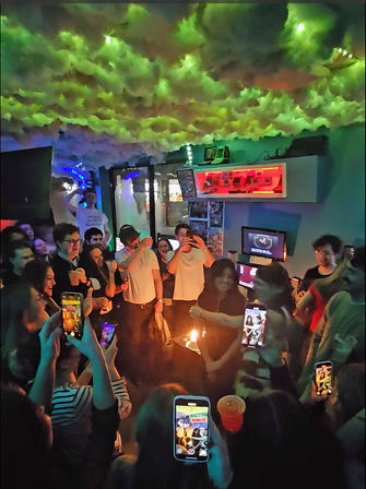 Vibrant urban nightlife scene: friends crowd a small bar for a birthday celebration, candles on a cake, phones recording under neon lights and a cloud-like ceiling installation.