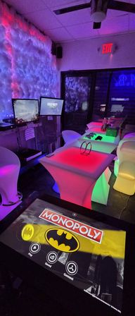 Neon-lit indoor gaming lounge with glowing RGB cocktail tables, touchscreen showing Batman Monopoly, dual monitors displaying Call of Duty, cloud-textured LED wall and modern bar stools