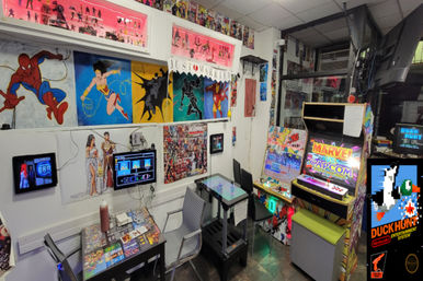 Colorful retro arcade corner with a Marvel vs. Capcom cabinet, Duck Hunt poster, comic-hero murals, a cocktail-style touchscreen table, chairs and collectible figures on shelves.