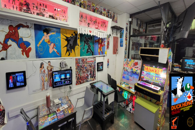 Colorful retro arcade corner with a Marvel vs. Capcom cabinet, Duck Hunt poster, comic-hero murals, a cocktail-style touchscreen table, chairs and collectible figures on shelves.