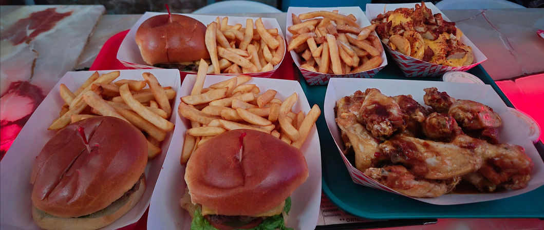 Diner-style spread with two cheeseburgers on brioche buns and fries, extra trays of golden fries and loaded potato rounds, and a basket of BBQ-glazed chicken wings on a tray
