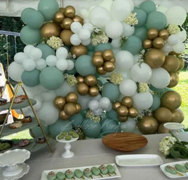 Mint green, white and metallic gold balloon wall with floral accents behind a dessert table featuring green macarons and serving trays at an outdoor party tent.
