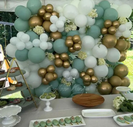 Mint green, white and metallic gold balloon wall with floral accents behind a dessert table featuring green macarons and serving trays at an outdoor party tent.