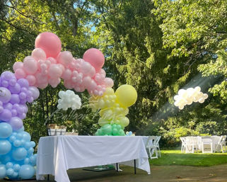 Pastel rainbow balloon arch framing a white-clothed table on a sunlit backyard lawn, white folding chairs set for an outdoor garden party.