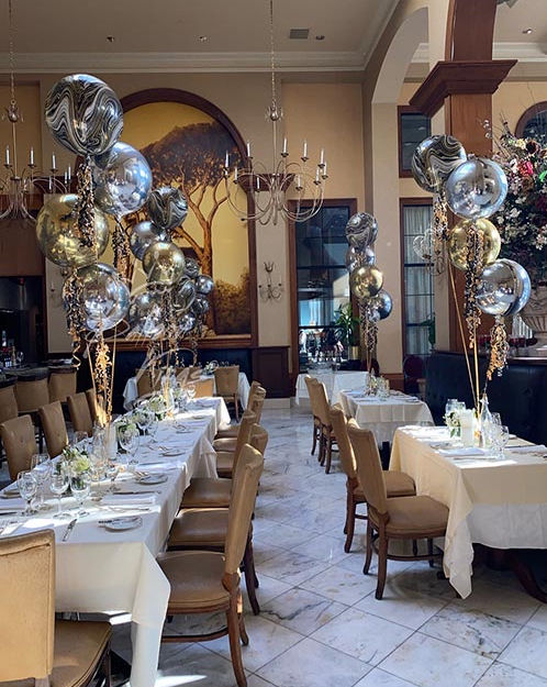 Elegant restaurant dining room set for a celebration with long white‑linen tables and place settings, tan leather chairs on marble floors, clusters of gold and silver metallic balloons, chandeliers and a large tree mural.