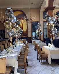 Elegant restaurant dining room set for a celebration with long white‑linen tables and place settings, tan leather chairs on marble floors, clusters of gold and silver metallic balloons, chandeliers and a large tree mural.