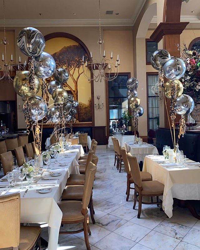 Elegant restaurant dining room set for a celebration with long white‑linen tables and place settings, tan leather chairs on marble floors, clusters of gold and silver metallic balloons, chandeliers and a large tree mural.