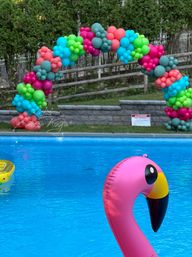 Pink inflatable flamingo floating in a bright blue backyard pool with a colorful teal, lime, coral and magenta balloon arch decorating the poolside for a summer pool party