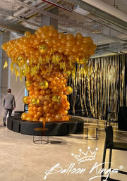 Oversized orange and metallic-gold balloon tree installation in a modern industrial indoor event space, surrounded by round black seating and a shimmering gold fringe backdrop.