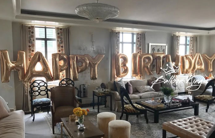 Spacious sunlit living room decorated for a birthday with large rose-gold "HAPPY BIRTHDAY" foil balloons across windows, plush sofas, coffee table and floral accents.