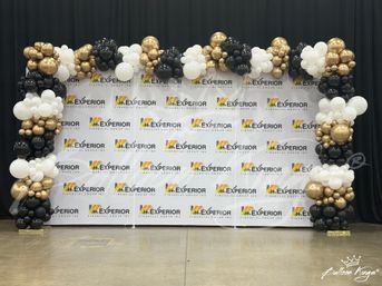 Festive black, white, and gold balloon arch framing a corporate step-and-repeat photo backdrop in an indoor event hall with polished concrete floor and black drapes.