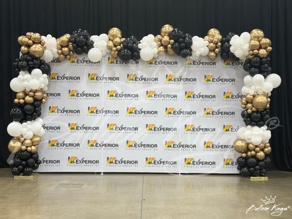 Festive black, white, and gold balloon arch framing a corporate step-and-repeat photo backdrop in an indoor event hall with polished concrete floor and black drapes.
