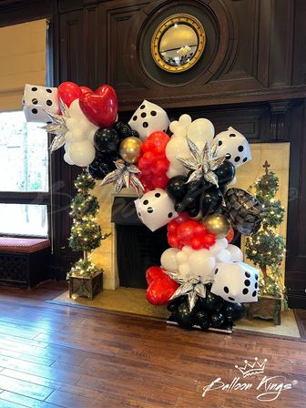 Casino-themed balloon garland with oversized dice, red heart balloons and silver star balloons in black, white, red and gold framing a fireplace in a wood-paneled indoor venue with lit topiary trees