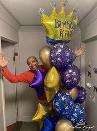 Person peeking from a doorway holding a large birthday balloon bouquet of gold, purple and blue foil stars and round balloons topped by a crown-shaped "Birthday King" balloon in an indoor hallway