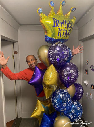 Person peeking from a doorway holding a large birthday balloon bouquet of gold, purple and blue foil stars and round balloons topped by a crown-shaped "Birthday King" balloon in an indoor hallway