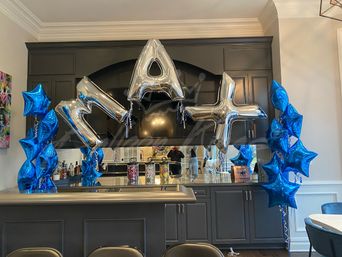 Modern gray home kitchen bar decorated for a celebration with oversized silver letter balloons and clusters of shiny blue star-shaped balloons framing a mirrored backsplash and countertop treats.