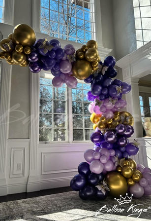 Purple, lavender and metallic gold balloon garland arch with butterfly accents framing tall paned windows in a bright, elegant white‑trimmed room — indoor event decor backdrop for weddings, parties or photo ops.