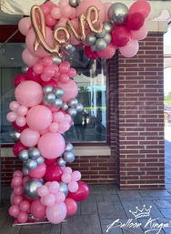 Cheerful pink and silver balloon column and arch at a brick storefront entrance, clustered pink balloons with metallic silver accents topped by a rose gold "love" script foil balloon