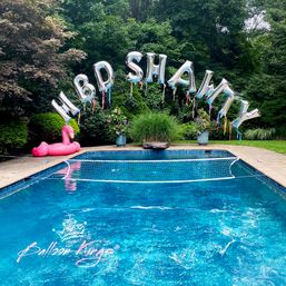 Backyard pool party setup: clear blue swimming pool with a net across the water, silver foil letter balloons arched overhead spelling a birthday message, a pink flamingo float on the deck, and lush green garden backdrop.