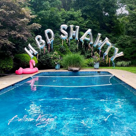 Backyard pool party setup: clear blue swimming pool with a net across the water, silver foil letter balloons arched overhead spelling a birthday message, a pink flamingo float on the deck, and lush green garden backdrop.
