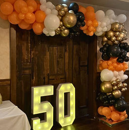Illuminated marquee 50 beside rustic wooden double doors, framed by orange, white, black and metallic gold balloon garland at an indoor party entrance.