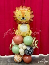 Smiling lion foil balloon perched atop a safari-themed balloon column with green, terracotta and cream latex balloons, zebra and giraffe print balloons and gold spiral accents against a red curtain backdrop.