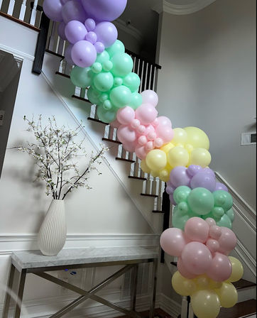 Playful pastel balloon garland in purple, mint, pink and yellow draped along a white staircase banister above a marble-top console table with a fluted vase of white blossom branches in a home foyer.