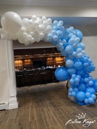 Blue and white balloon garland arch framing an interior doorway to a home bar with dark wood cabinets, bar stools and wood-look tile flooring — indoor party decoration.