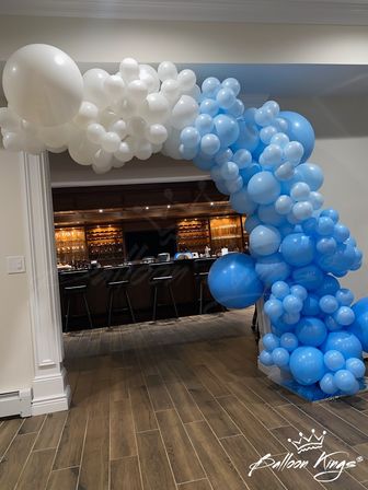 Blue and white balloon garland arch framing an interior doorway to a home bar with dark wood cabinets, bar stools and wood-look tile flooring — indoor party decoration.