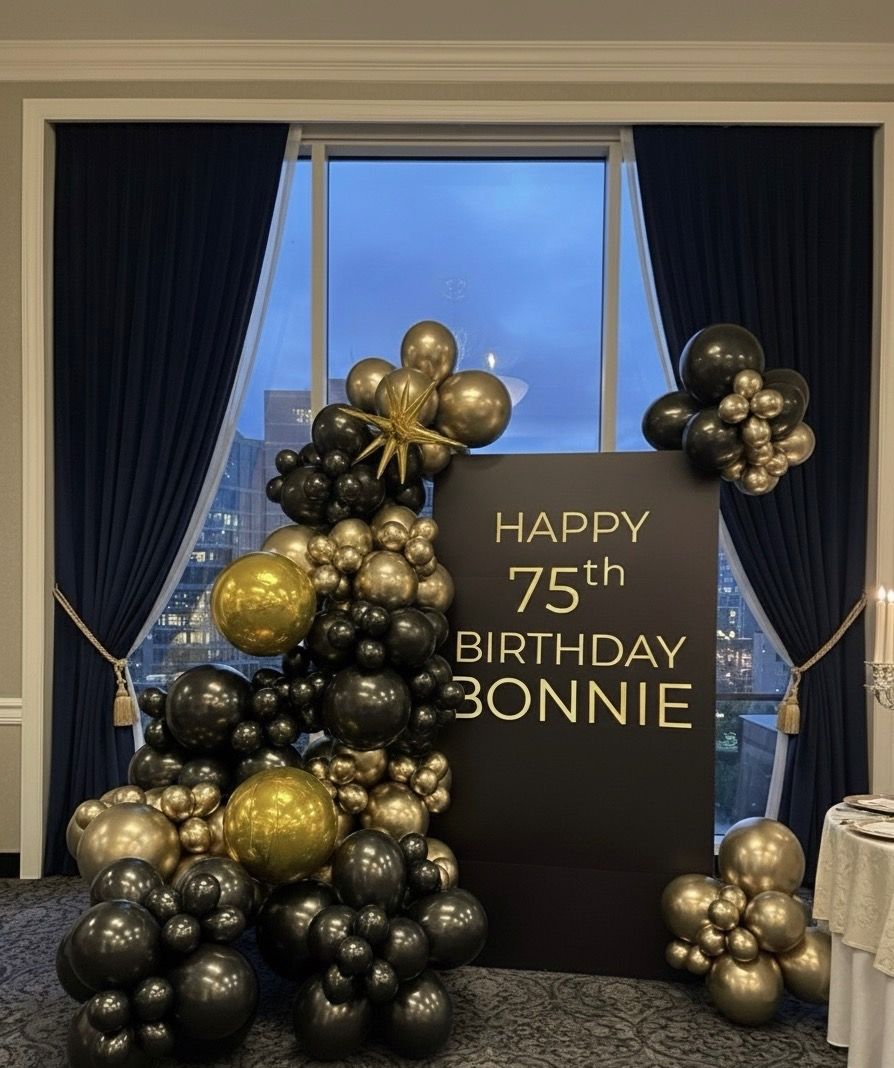Elegant black-and-gold balloon garland framing a matte black sign reading HAPPY 75th BIRTHDAY in a banquet room with navy drapes and a city skyline visible through a tall window.