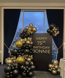 Elegant black-and-gold balloon garland framing a matte black sign reading HAPPY 75th BIRTHDAY in a banquet room with navy drapes and a city skyline visible through a tall window.