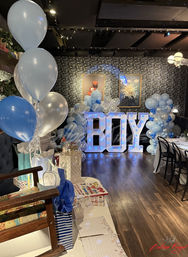 Blue-and-white baby shower setup in an indoor venue with giant illuminated 'BOY' letters, balloon garlands and clusters, and a decorated gift table.