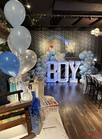 Blue-and-white baby shower setup in an indoor venue with giant illuminated 'BOY' letters, balloon garlands and clusters, and a decorated gift table.