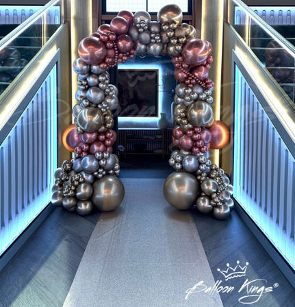 Shimmering metallic rose-gold and silver balloon archway framing a modern indoor staircase entrance with blue LED-lit railings, stylish event decor