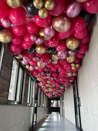 Indoor corridor with a balloon ceiling installation — dense canopy of fuchsia, metallic gold, rose-gold, black and mauve balloons lining the hallway above windows and glass doors