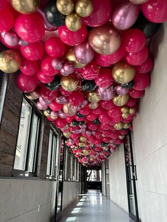 Indoor corridor with a balloon ceiling installation — dense canopy of fuchsia, metallic gold, rose-gold, black and mauve balloons lining the hallway above windows and glass doors