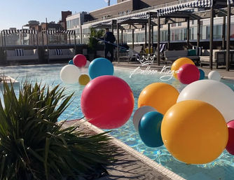 Playful oversized colorful balloons floating in a sunlit urban rooftop pool with cabanas, lounge seating and city buildings in the background.