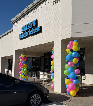 Cheerful white brick sandwich shop storefront in a strip mall, entrance flanked by colorful balloon columns, parked car in front and clear blue sky