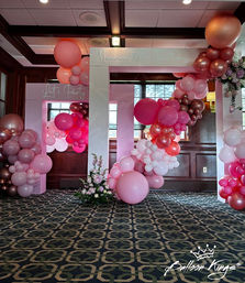 Festive indoor birthday photo frame with pink and rose‑gold balloon arch, oversized balloons and floral accents in a wood‑paneled event room with patterned carpet.