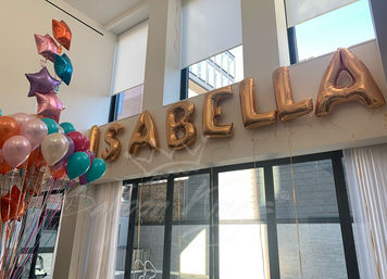 Rose-gold foil letter balloons spelling "ISABELLA" hung near tall windows in a bright modern room, surrounded by teal, pink, purple and orange helium balloons and star-shaped foil balloons — festive indoor party decor.